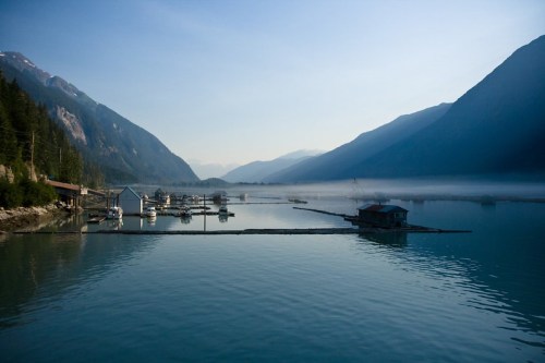 Serene lake surrounded by mountains, with boats docked and mist rising in the early morning light.