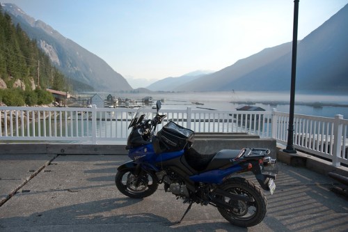 A blue motorcycle parked by a scenic waterfront with mountains and mist in the background.