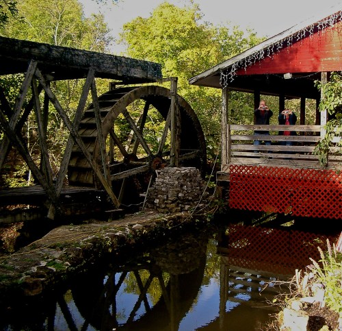 A rustic watermill beside a calm stream, with a red building and lush greenery in the background.