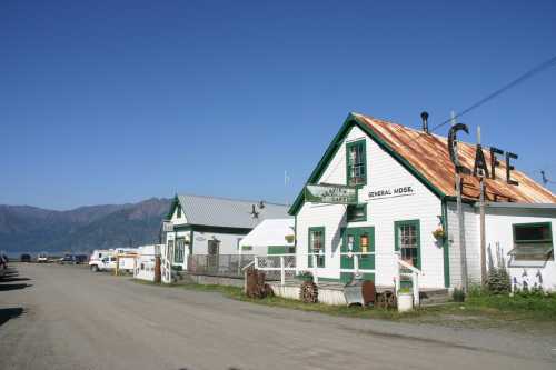 A quaint café and general store with green trim, set against a clear blue sky and mountains in the background.