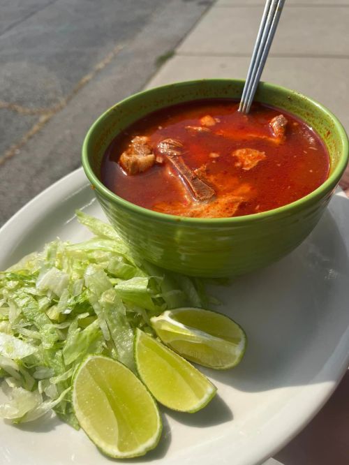 A green bowl of red soup with meat, served with shredded lettuce and lime wedges on a plate.
