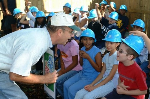 A group of children in blue hard hats listens attentively to an adult speaking animatedly in a playful outdoor setting.