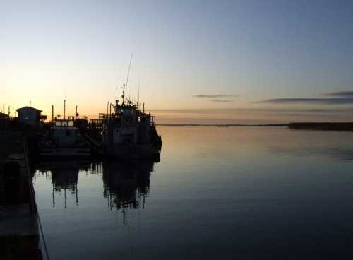 Silhouette of boats at a calm river during sunset, with soft colors reflecting on the water's surface.