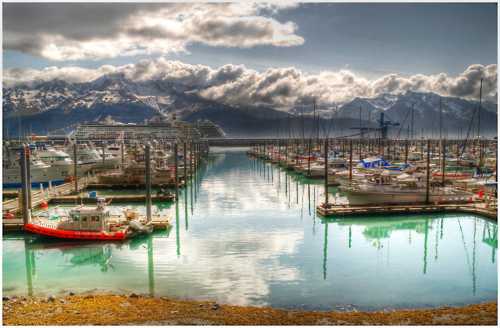 A serene marina filled with boats, reflecting mountains and clouds in calm water under a bright sky.