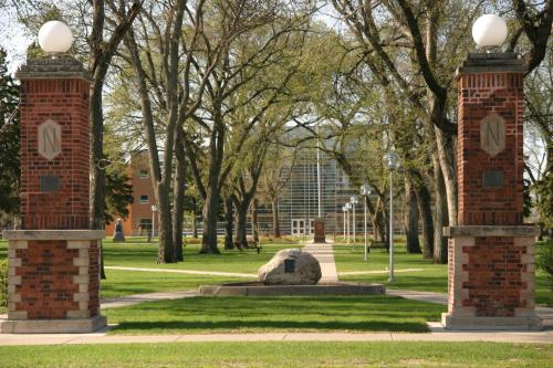 A campus entrance with brick pillars, trees, and a building in the background, set in a green park-like area.