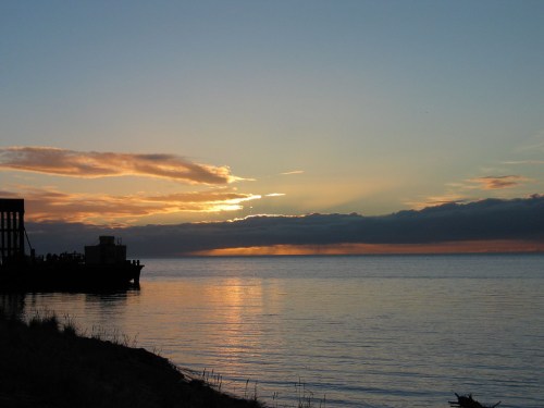 A serene sunset over calm waters, with silhouettes of a pier and clouds reflecting warm colors in the sky.