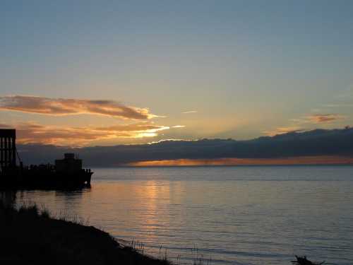 A serene sunset over calm waters, with silhouettes of a pier and clouds reflecting warm colors in the sky.