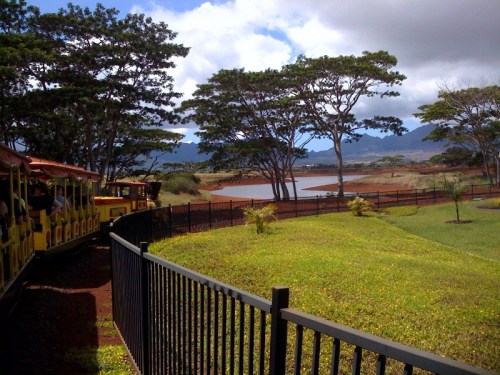 A scenic view of a train passing by a lake surrounded by trees and a black fence under a partly cloudy sky.