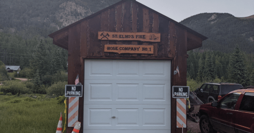 A rustic firehouse with a wooden sign reading "St. Elmo's Fire Hose Company No. 1" and "No Parking" signs outside.