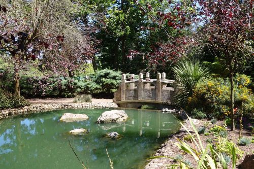 A serene garden scene featuring a stone bridge over a green pond, surrounded by lush trees and colorful flowers.