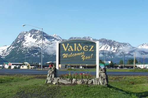 A welcome sign for Valdez, Alaska, with snow-capped mountains in the background and a clear blue sky.