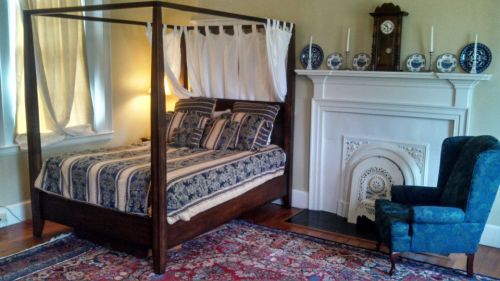A cozy bedroom featuring a four-poster bed, a fireplace, and a blue armchair, with decorative plates on the mantel.
