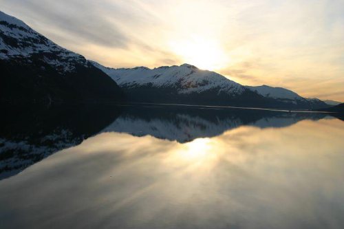 Sunset over a calm lake, reflecting snow-capped mountains and a colorful sky.