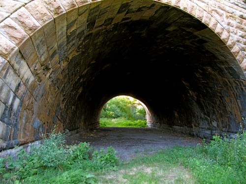 A stone archway tunnel with greenery visible at both ends, leading into a bright, natural landscape.