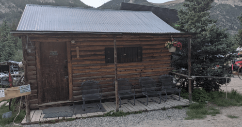 A rustic wooden cabin with a metal roof, featuring a porch and chairs, surrounded by trees and mountains.