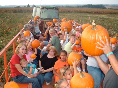 A group of people on a hayride, holding pumpkins and smiling, surrounded by a pumpkin patch and fields.