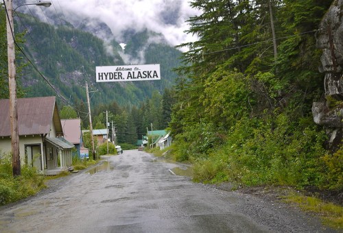 A scenic view of a road leading into Hyder, Alaska, with a welcome sign and lush green mountains in the background.