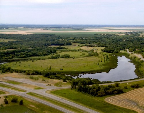 Aerial view of lush green fields, a winding river, and a mix of trees and open land under a clear sky.