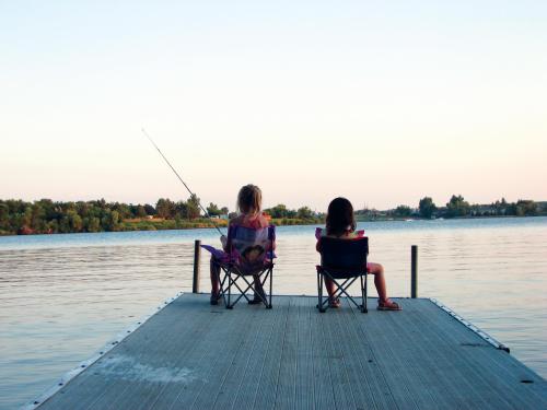 Two children sit on a dock, fishing and enjoying the calm water and sunset in the background.
