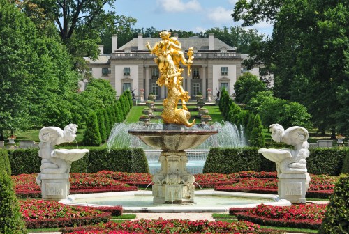 A grand fountain with a golden statue, surrounded by lush gardens and a mansion in the background.