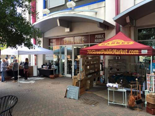Entrance to a public market with tents and vendors outside, showcasing various goods and a lively atmosphere.