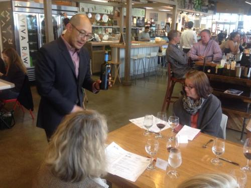 A man presents a wine bottle to two seated women at a restaurant table, with glasses and menus in front of them.