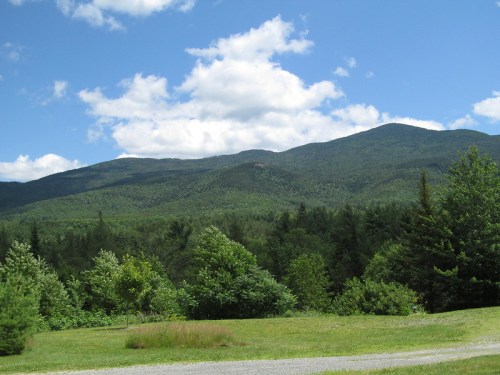 Lush green mountains under a blue sky with fluffy clouds, surrounded by trees and open grassy areas.