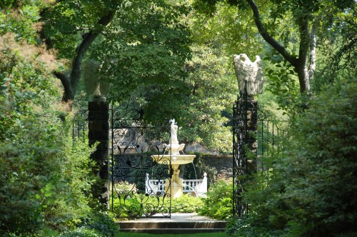 A serene garden entrance with a wrought iron gate, flanked by statues, leading to a fountain surrounded by lush greenery.
