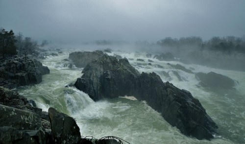 A misty river flows over rocky terrain, surrounded by fog and trees, creating a dramatic, moody landscape.