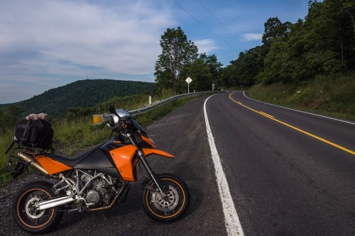 A parked orange motorcycle beside a winding road, with green hills and a blue sky in the background.