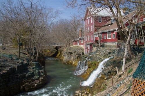 A red building beside a flowing river with small waterfalls, surrounded by bare trees and a clear blue sky.