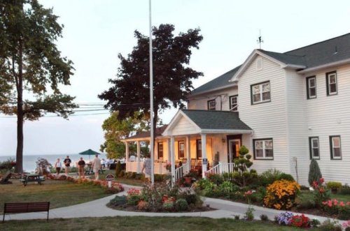 A charming white house with a porch, surrounded by colorful gardens, near a lake at sunset. People gather outside.