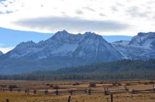 Snow-capped mountains rise above a grassy meadow, with a wooden fence in the foreground under a blue sky.