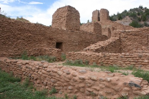 Ancient adobe ruins with stone walls and towers, surrounded by greenery under a blue sky.