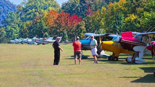 Three people chat on a grassy field beside a row of colorful small airplanes, with trees displaying autumn foliage in the background.