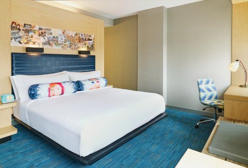 A modern hotel room featuring a large bed, colorful decor, a desk, and a stylish chair on a blue carpet.