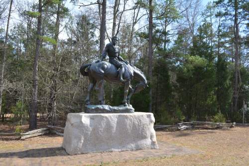 A bronze statue of a mounted soldier on a horse, set in a wooded area with trees in the background.