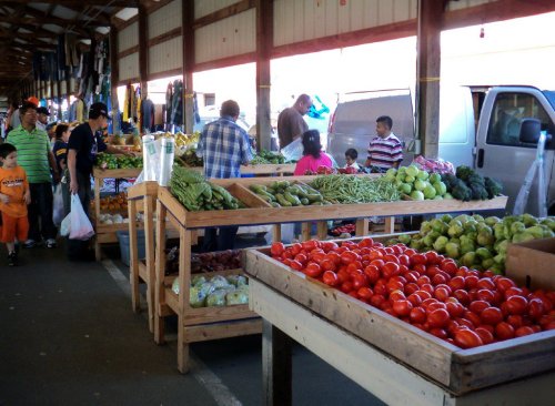 A bustling farmers market with various fresh vegetables and fruits displayed on wooden tables, and people shopping.