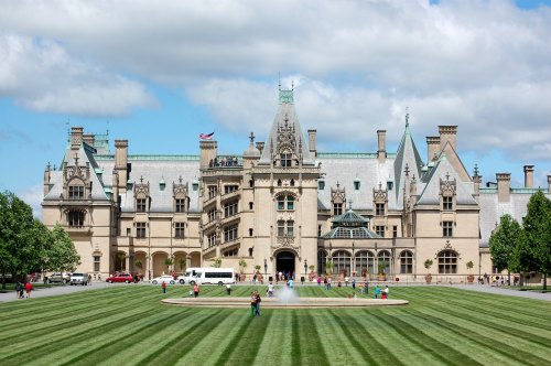 A grand mansion with intricate architecture, surrounded by manicured lawns and a fountain, under a partly cloudy sky.