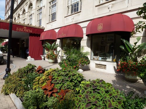 A storefront with red awnings, lush plants, and colorful flowers in front of a building with elegant architecture.