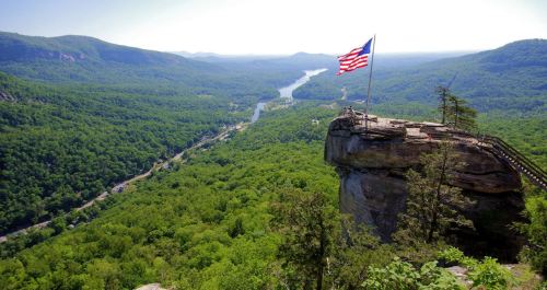A scenic view from a rocky overlook with a U.S. flag, surrounded by lush green mountains and a winding river below.