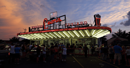A bustling ice cream stand at sunset, with a long line of customers and bright neon signs.