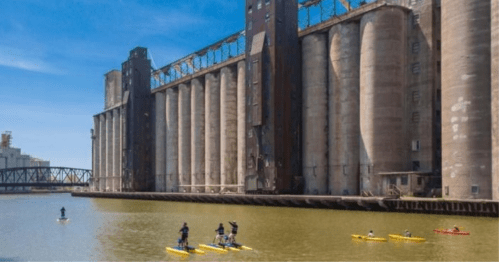 Kayakers paddle in front of large industrial silos along a calm river under a clear blue sky.