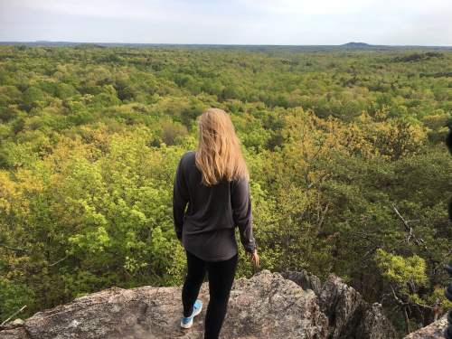 A person with long hair stands on a rocky ledge, overlooking a lush green landscape and distant hills.