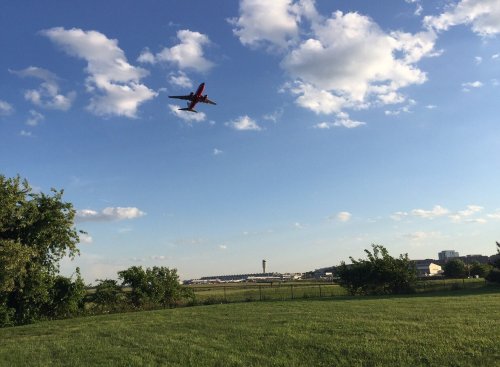 An airplane flies overhead against a blue sky with clouds, near an airport with a control tower in the background.