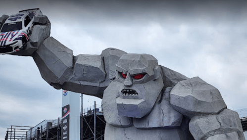 A large stone statue of a giant holding a race car, with a fierce expression, set against a cloudy sky.