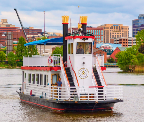 A colorful riverboat with a tall smokestack navigates a calm waterway, surrounded by city buildings in the background.