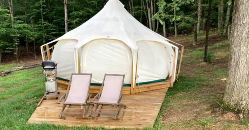 A white tent on a wooden deck in a forested area, with two lounge chairs and a grill nearby.