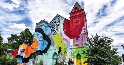 Colorful mural on a historic building under a blue sky with clouds, surrounded by greenery.