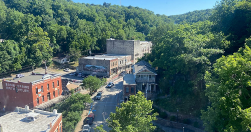 Aerial view of a small town surrounded by lush greenery, featuring historic buildings and a winding road.